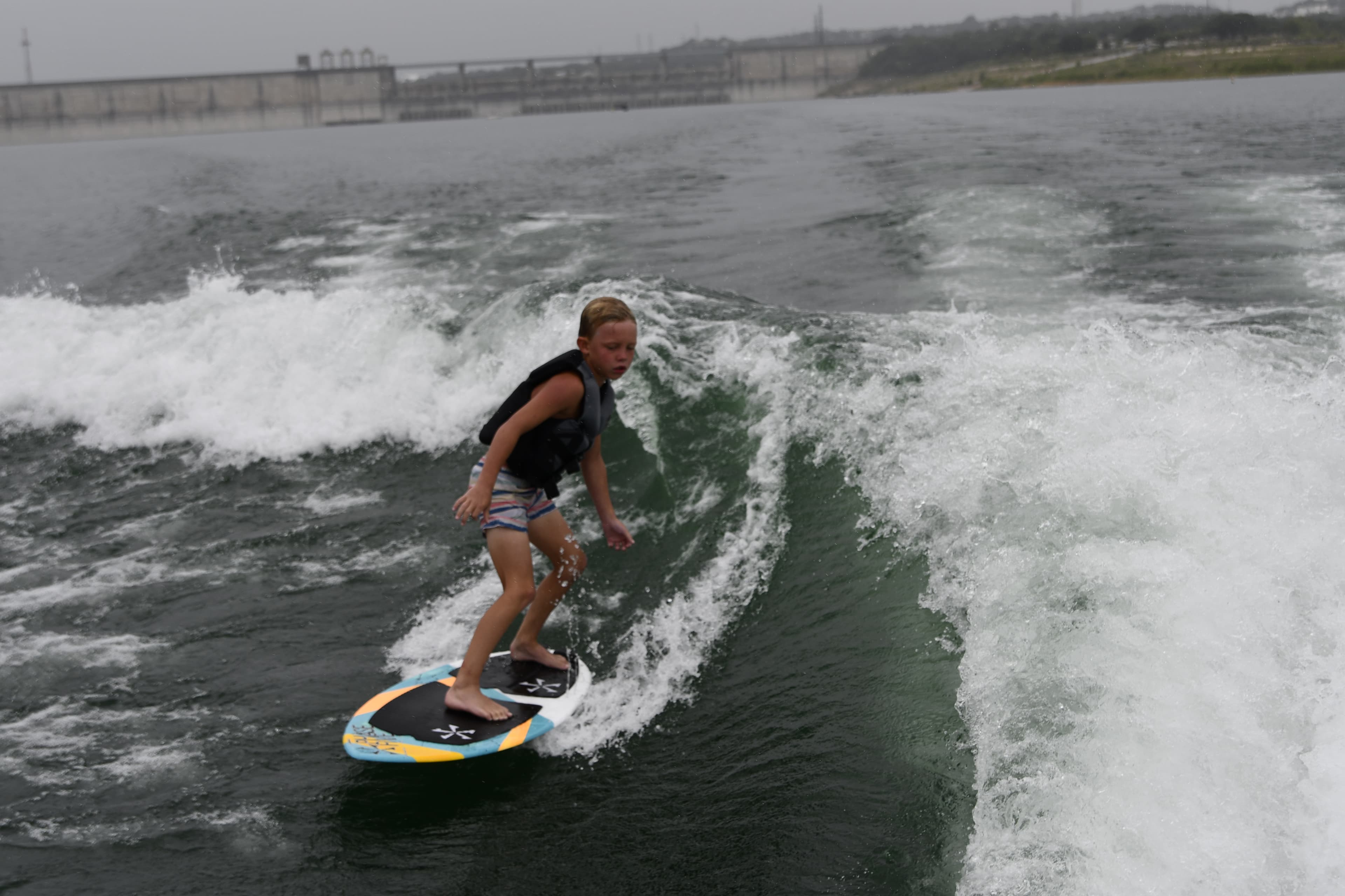 A wakesurfer carving a clean wake on Lake Travis with the Mansfield Dam visible in the background on a clear Texas morning.