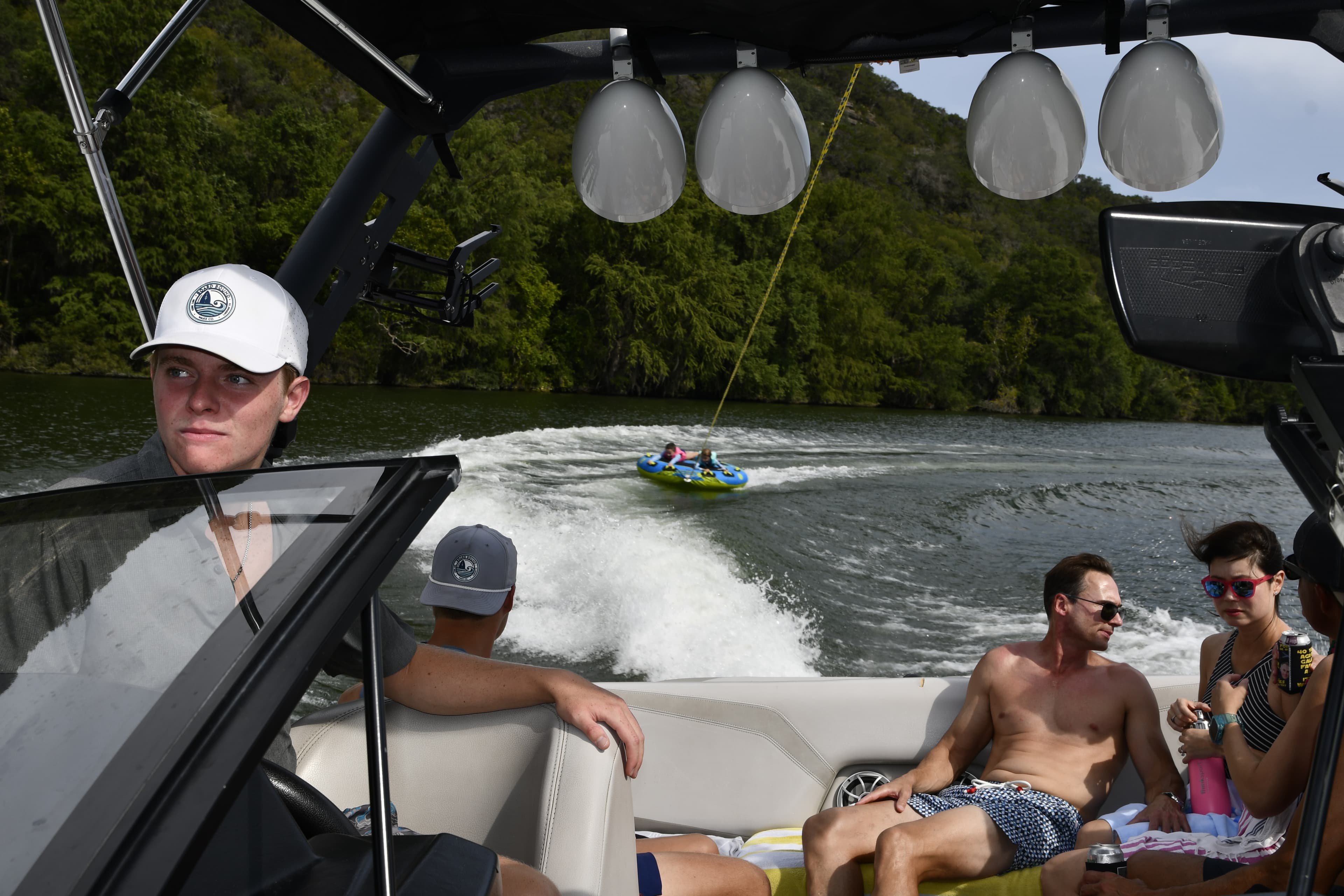 Friends relaxing on a Board Bound Wake Co boat on a sunny Texas hill country lake with a tuber visible in the background.