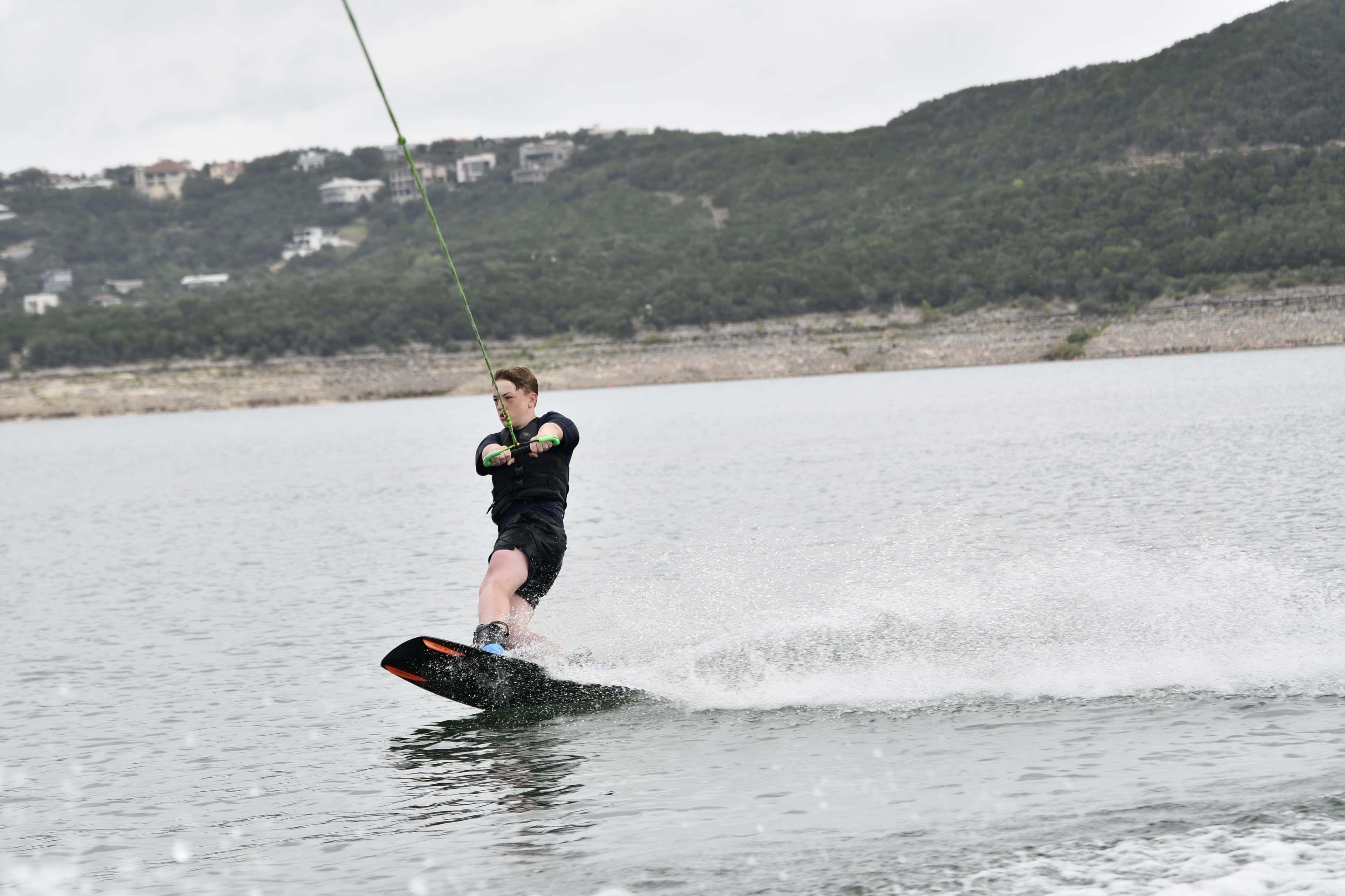 A wakesurfer carving a clean wave behind a wakeboat on Lake Travis with the Mansfield Dam shoreline in the background
